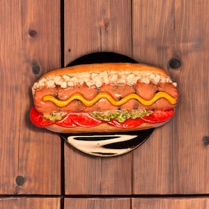 Puede incluir: Primer plano de un perrito caliente sobre un fondo de madera. El perrito caliente está cubierto con mostaza, salsa y cebollas. Está sobre un plato negro.