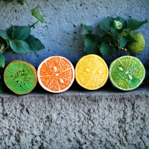 May include: Four halved citrus fruits arranged in a row on a gray textured surface. From left to right, the fruits are a green kiwi, an orange orange, a yellow lemon, and a green lime.
