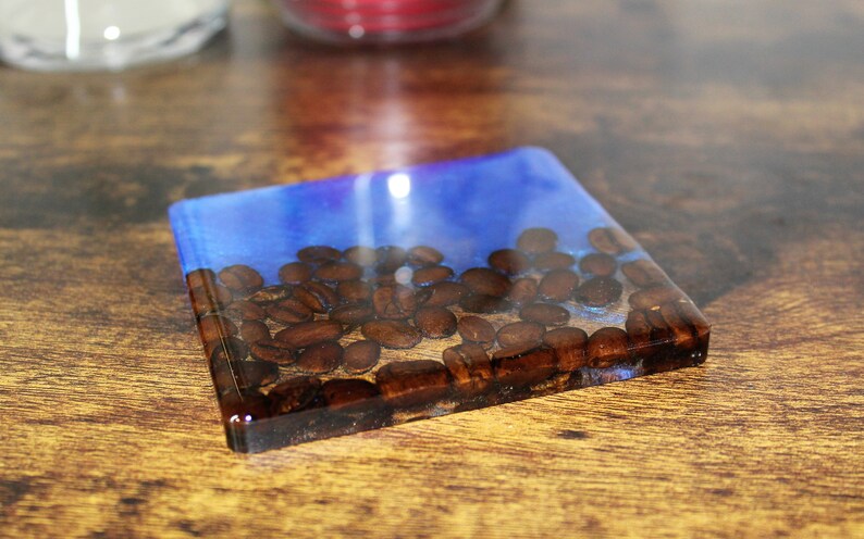 May include: A square blue and clear resin coaster with coffee beans embedded inside. The coaster is sitting on a wooden surface.