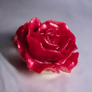 May include: A vibrant red rose-shaped candle. The candle's petals are detailed and glossy, with a slight translucent quality. The rose sits on a clear, circular base, suggesting it is a decorative item.