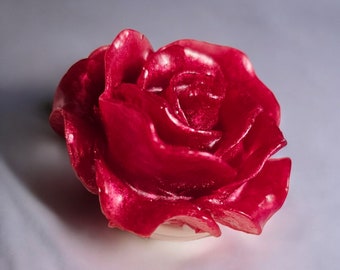 May include: A vibrant red rose-shaped candle. The candle's petals are detailed and glossy, with a slight translucent quality. The rose sits on a clear, circular base, suggesting it is a decorative item.