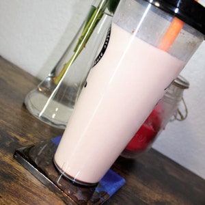 May include: A pink milk tea drink with a clear plastic cup and a black lid. The drink is sitting on a brown and blue resin coaster.