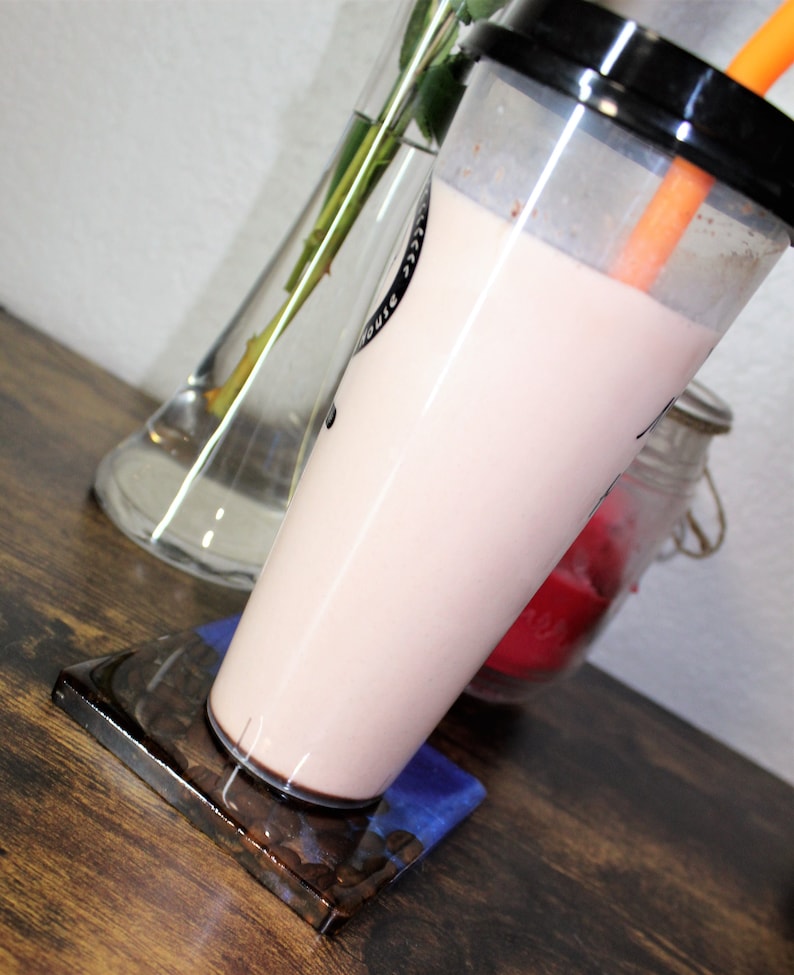 May include: A pink milk tea drink with a clear plastic cup and a black lid. The drink is sitting on a brown and blue resin coaster.