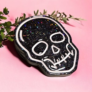 May include: Black glitter skull with a white outline. The skull has a detailed design with a glittery finish.