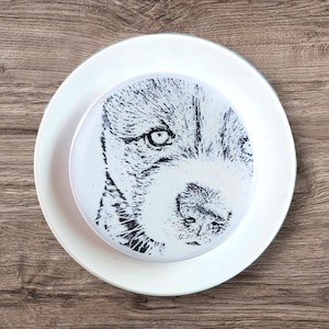 May include: A white ceramic plate with a black and white image of a dog's face printed on it. The dog's eye is visible and the image is centered on the plate.