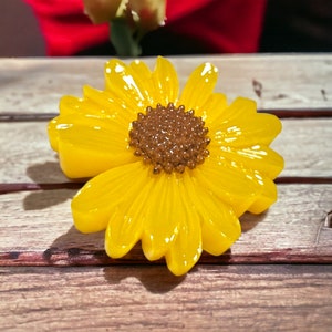 May include: A yellow resin sunflower brooch with a brown center. The brooch is on a wooden surface.