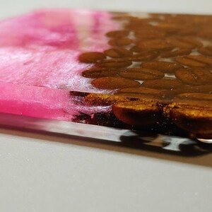 May include: A pink and brown resin coaster with a design of coffee beans. The coaster is made of clear resin with a pink layer on top and a layer of brown coffee beans on the bottom.