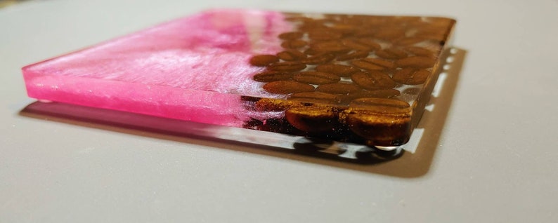 May include: A pink and brown resin coaster with a design of coffee beans. The coaster is made of clear resin with a pink layer on top and a layer of brown coffee beans on the bottom.