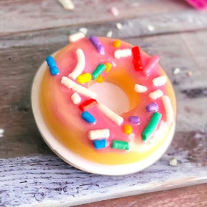 May include: A pink frosted donut with colorful sprinkles on a white plate. The donut is on a wooden surface.