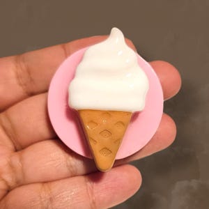 May include: A pink phone grip with a white ice cream cone design. The ice cream is white and the cone is brown.