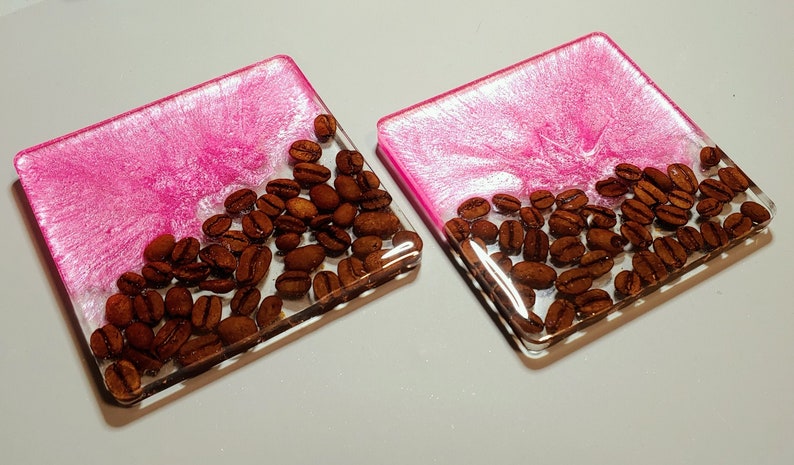 May include: Two pink and white resin coasters with a layer of coffee beans embedded in the resin.