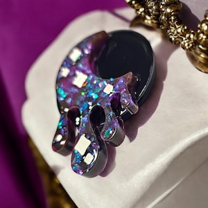 Puede incluir: Un colgante de resina morado y azul con forma de luna creciente con purpurina blanca incrustada en la resina.