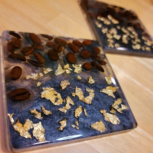 May include: Two square coasters with a black and white resin base. The coasters are decorated with gold leaf and coffee beans.