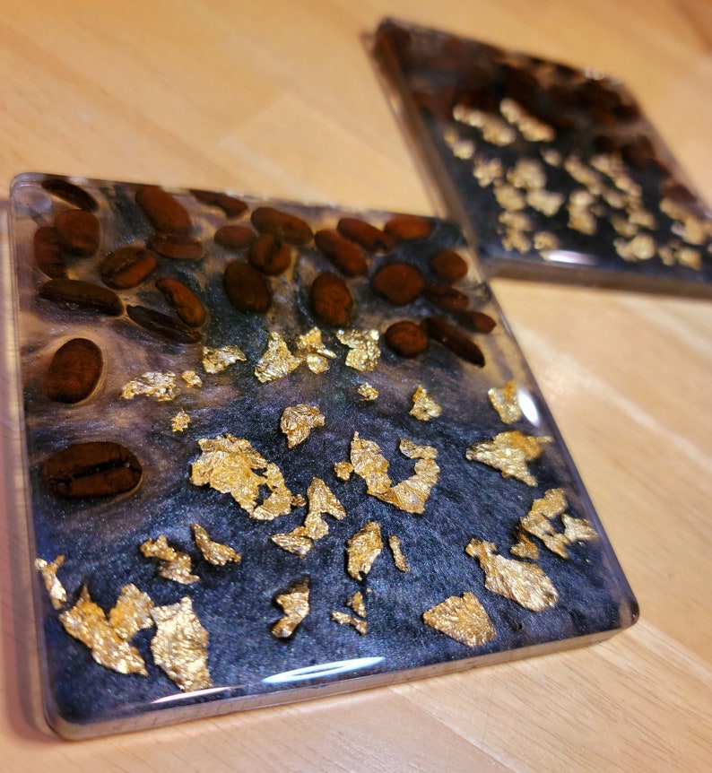 May include: Two square coasters with a black and white resin base. The coasters are decorated with gold leaf and coffee beans.