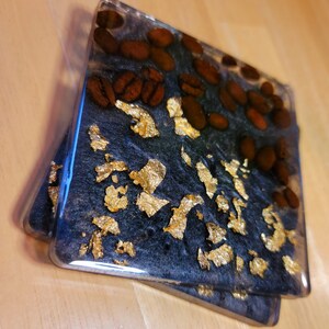 May include: Two square black coasters with gold leaf and brown coffee beans embedded in the resin. The coasters are clear on the edges.