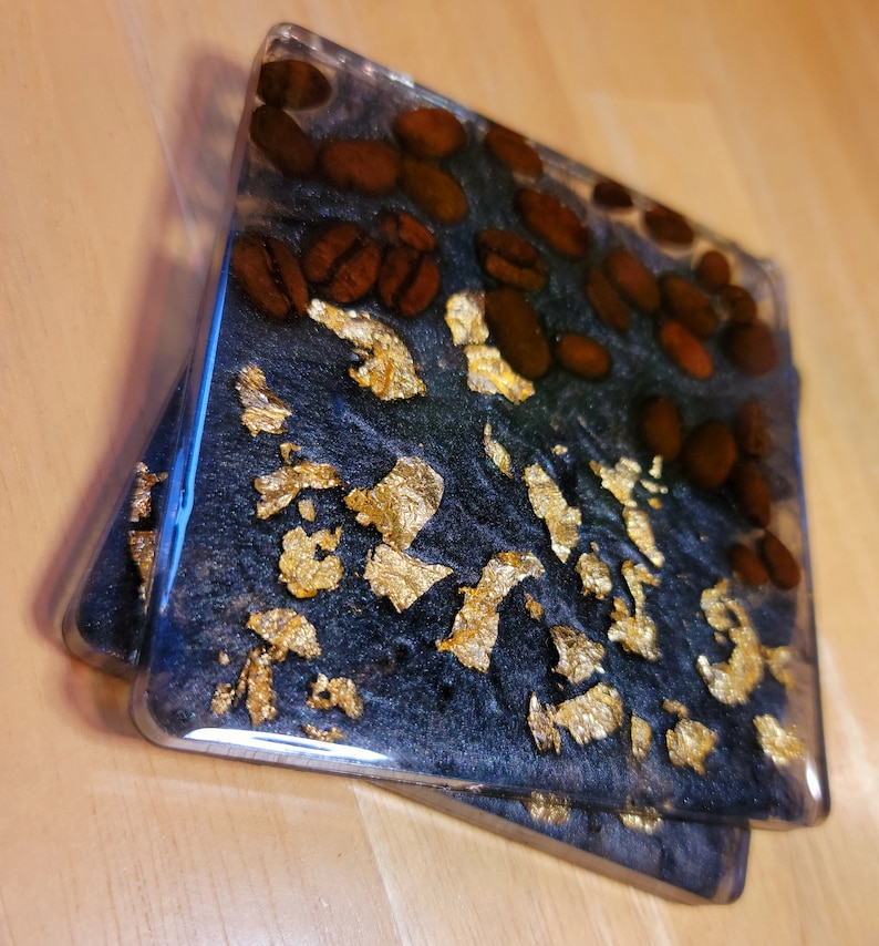 May include: Two square black coasters with gold leaf and brown coffee beans embedded in the resin. The coasters are clear on the edges.