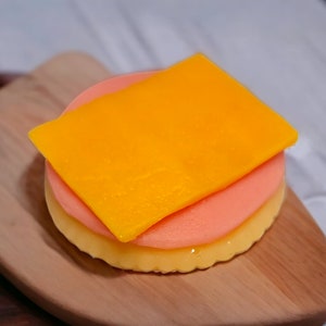 Pode incluir: Uma sobremesa em camadas sobre uma tábua de cortar de madeira. A sobremesa é feita de três camadas: uma camada amarela, uma camada rosa e uma camada amarela no topo.