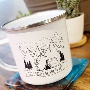 May include: White enamel camping mug with a black and white illustration of mountains, a tent, a campfire, and trees. The mug has a silver rim and the text "This must be the place" on a banner.
