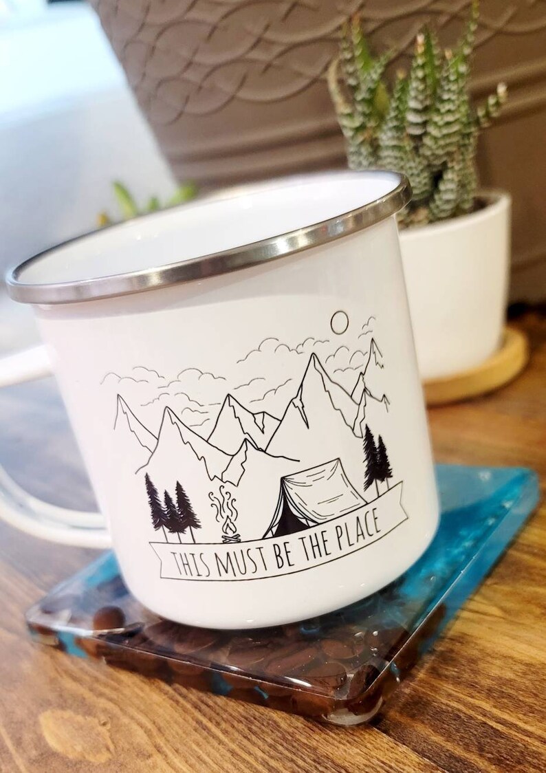 May include: White enamel camping mug with a black and white illustration of mountains, a tent, a campfire, and trees. The mug has a silver rim and the text "This must be the place" on a banner.
