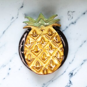 Puede incluir: Un plato dorado en forma de piña con la parte superior verde, sobre una superficie de mármol blanco y negro.