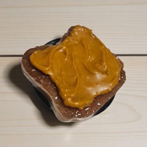 May include: A novelty phone grip featuring a slice of toast with peanut butter. The toast is brown, and the peanut butter is a light brown color. The phone grip is black.