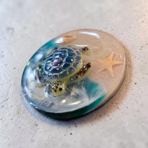 Pode incluir: Um porta-copos circular de resina com uma tartaruga marinha tridimensional de carapaça azul e amarela a nadar em resina transparente. O design inclui uma base bege arenosa, uma pequena estrela-do-mar clara e ondas turquesa e brancas.