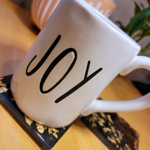 May include: White ceramic mug with the word "JOY" written in black script. The mug is sitting on a black and gold coaster.