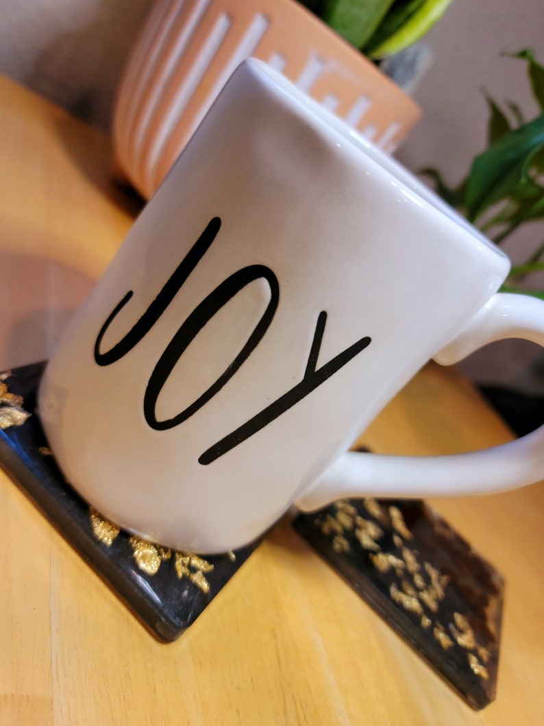 May include: White ceramic mug with the word "JOY" written in black script. The mug is sitting on a black and gold coaster.