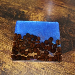 May include: A square blue and brown resin coaster with a design of coffee beans.