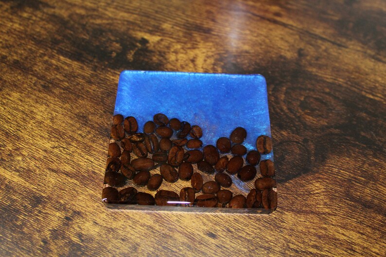May include: A square blue and brown resin coaster with a design of coffee beans.