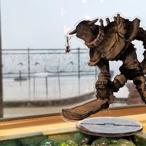Può includere: Una statuetta marrone e grigia di una creatura con una grande testa e una spada in mano. La statuetta è su un supporto in plastica trasparente con una base nera.