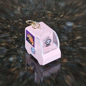 Peut inclure: Un porte-clés en plastique rose en forme de fourgon avec une fenêtre en plastique transparent. Le fourgon a un autocollant de personnage de dessin animé sur le côté et une petite figurine sur le siège conducteur.