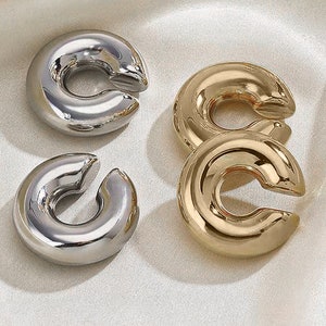 Puede incluir: Cuatro pendientes de aro, dos plateados y dos dorados, se muestran sobre una superficie blanca. Los pendientes tienen forma de círculo con un hueco en el medio.