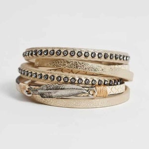 May include: A multi-layered bracelet with a gold and cream leather design. The bracelet features a small silver feather charm and black rhinestones.