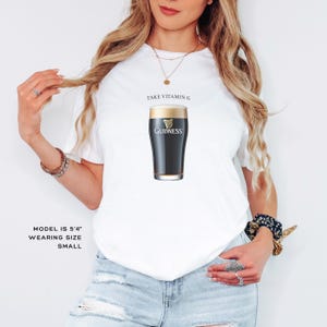 Puede incluir: Camiseta blanca con el texto "Take Vitamin G" y un gr&aacute;fico de un vaso de cerveza Guinness. La modelo lleva una talla peque&ntilde;a.
