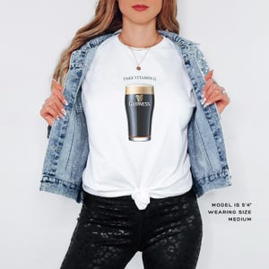 Puede incluir: Camiseta blanca con un gr&aacute;fico de vaso de pinta Guinness negro y dorado y el texto "Take Vitamin G".