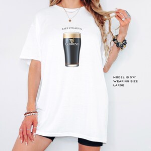 Puede incluir: Camiseta blanca con el texto "Take Vitamin G" y un gr&aacute;fico de un vaso de cerveza Guinness. La modelo lleva una talla grande.