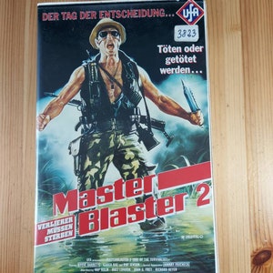 Könnte beinhalten: Ein VHS-Videokassetten-Cover für den Film "Master Blaster 2: Day of the Survivalist". Das Cover zeigt einen Mann in Tarnkleidung, der eine Waffe und ein Messer hält, mit dem Titel "Master Blaster 2" in rot-weißer Schrift. Der Text "Töten oder getötet werden..." ist ebenfalls auf dem Cover zu sehen.