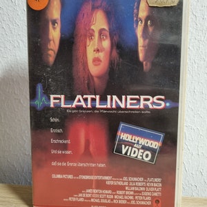 Könnte beinhalten: Ein VHS-Videokassetten-Etui für den Film "Flatliners" mit Julia Roberts, Kiefer Sutherland und Kevin Bacon. Das Etui ist rot und schwarz mit einem Hollywood on Video-Logo. Der Text auf dem Etui lautet "Es gibt Grenzen, die man nicht überschreiten sollte."