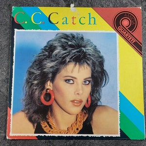 Könnte beinhalten: Ein Vinyl-Plattencover mit einer Frau mit blonden Haaren, die große rote Ohrringe und eine goldene Kettenkette trägt. Das Cover ist gelb mit blauen, grünen und roten Streifen. Der Text "C.C. Catch" ist in Rot gedruckt und der Text "AMIGA QUARTETT" ist in Schwarz gedruckt.