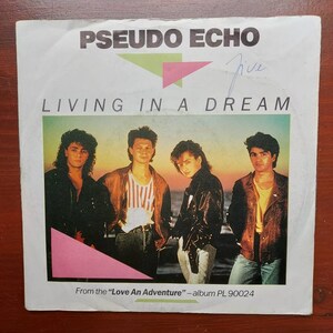 Könnte beinhalten: Eine 45-RPM-Vinyl-Single von Pseudo Echo für den Song "Living in a Dream". Die Platte stammt aus dem Album "Love An Adventure" und trägt die Albumnummer PL 90024, die auf dem Label gedruckt ist.
