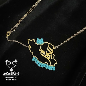 May include: A gold-coloured necklace with a pendant shaped like a country's map, embellished with turquoise beads and calligraphy. The necklace is suspended from a fine chain against a dark backdrop. The brand name "artsiAFRA" is visible.