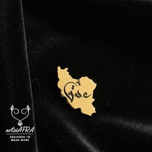 Könnte beinhalten: Eine goldfarbene Anstecknadel in Form eines Landes mit einem schwarzen Schriftzug. Die Anstecknadel befindet sich auf einer schwarzen Samtoberfläche. Der Text "artsiAFRA DESIGNED TO MEAN MORE" befindet sich in der unteren linken Ecke.