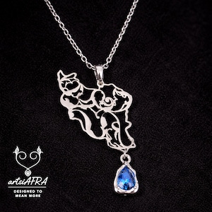 May include: A silver pendant necklace featuring a detailed cutout design of a map, with a teardrop-shaped blue gemstone. The necklace hangs on a delicate chain. The logo "artsiAFRA" and the text "DESIGNED TO MEAN MORE" are visible.