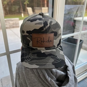 May include: A camouflage baseball cap with a brown leather patch that says "Rohda" in cursive.