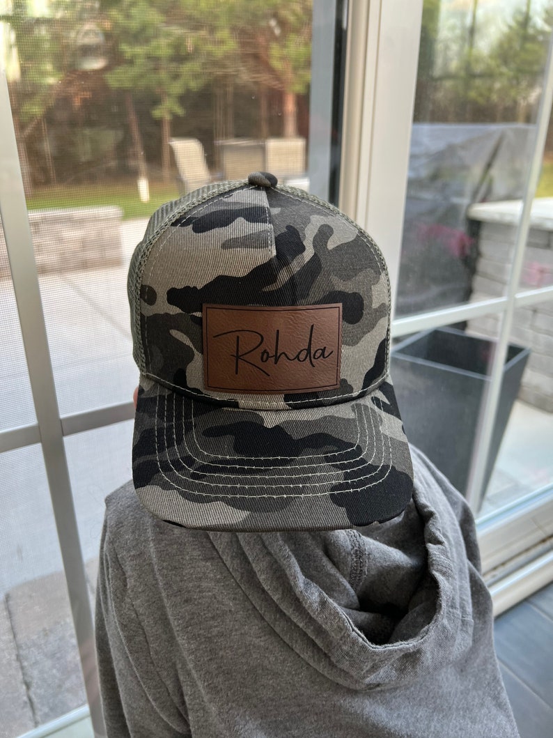 May include: A camouflage baseball cap with a brown leather patch that says "Rohda" in cursive.