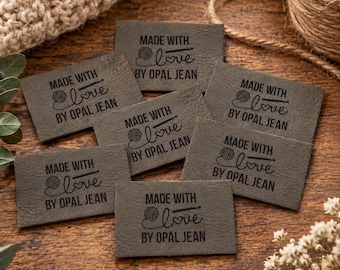 May include: A collection of rectangular dark gray labels with a textured surface. Each label features the black printed text MADE WITH love BY OPAL JEAN, accompanied by a small graphic of a ball of yarn and a heart symbol.