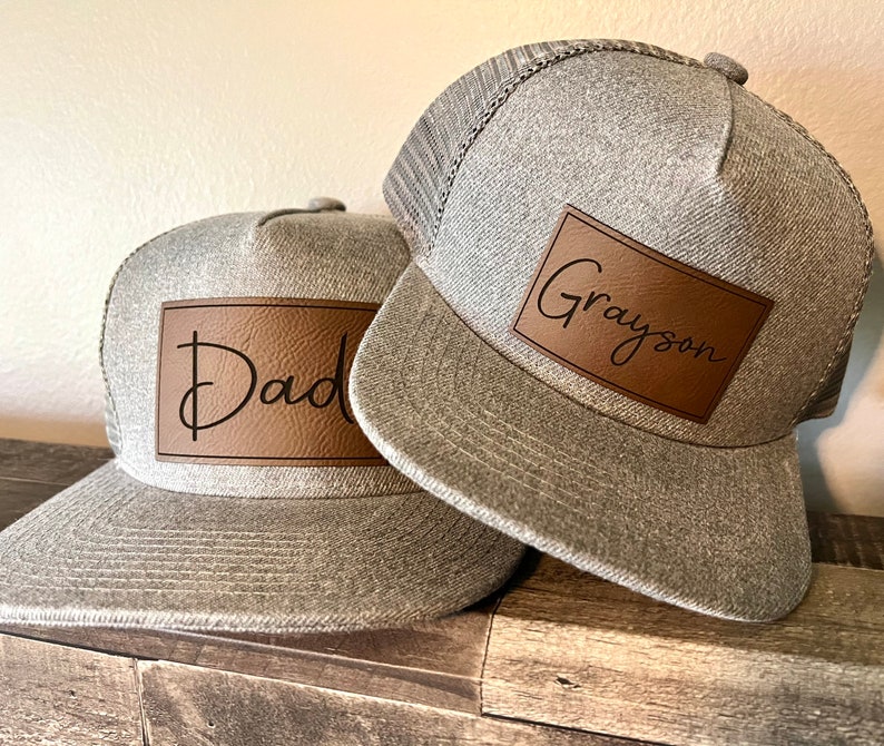 May include: Two gray baseball caps with brown leather patches. The patch on the left cap says "Dad" and the patch on the right cap says "Grayson".