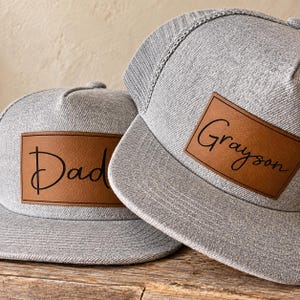 May include: Two heather grey baseball caps with flat brims and mesh backs. Each cap features a rectangular brown faux leather patch on the front. The left cap has the word Dad printed in black script, and the right cap has the name Grayson in black.