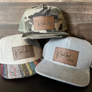 May include: Three grey and camo trucker hats with brown leather patches. The patches have the names "Rohda", "Kaislee", and "Kailor MAE" written in black script.